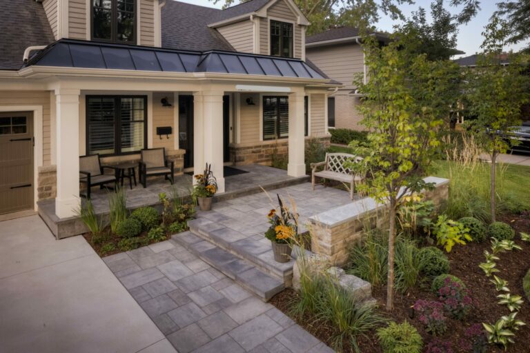 A two-story house with a spacious porch and garden. Stone path and steps are surrounded by plants and outdoor seating.