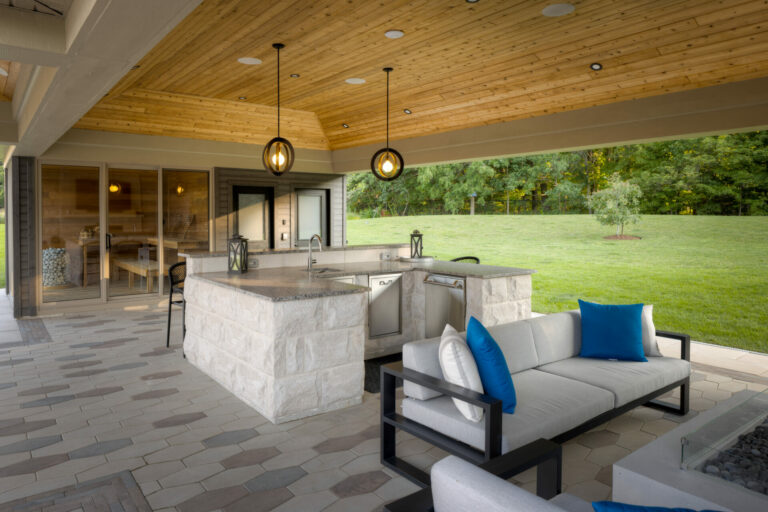 Stylish outdoor kitchen with stone countertops, modern lighting, and seating area. Glass doors lead to indoor dining; lush green lawn surrounds space.