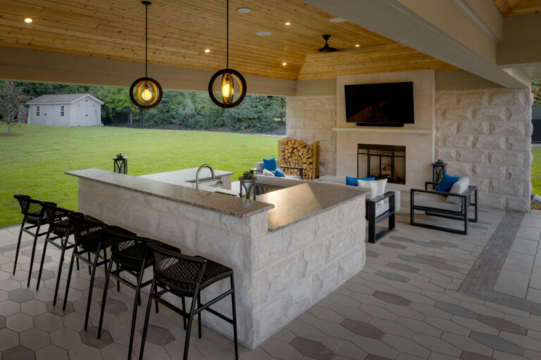 Outdoor living space features a covered patio with kitchen, bar seating, fireplace, TV, and seating area. Lawn and trees visible in background.