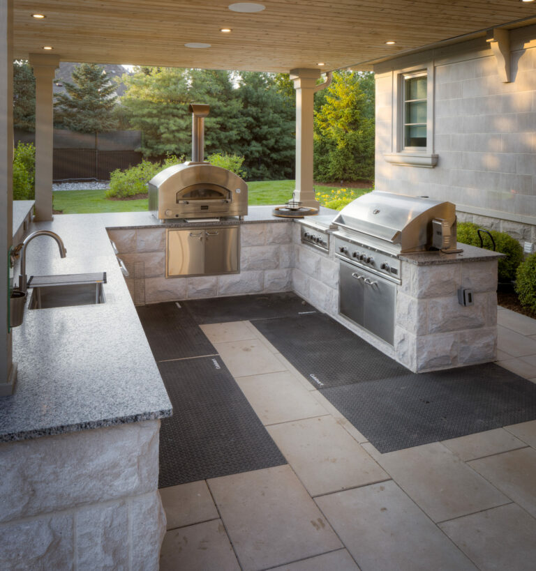 Outdoor kitchen with stone countertops, pizza oven, grill, and sink. Surrounded by trees and shrubs, under a wooden ceiling. Cozy backyard setting.