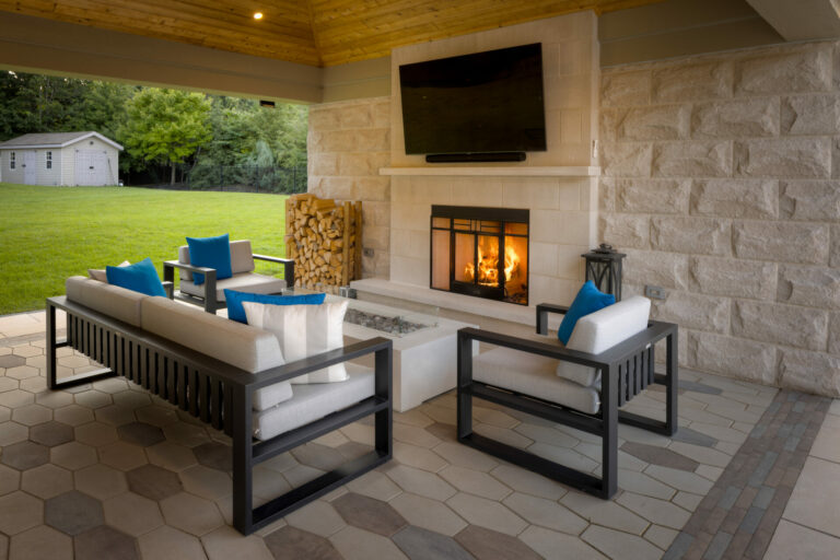 Modern outdoor living space with fireplace, comfortable seating, and TV. Features geometric tiles, stacked firewood, and serene backyard view with lush greenery.