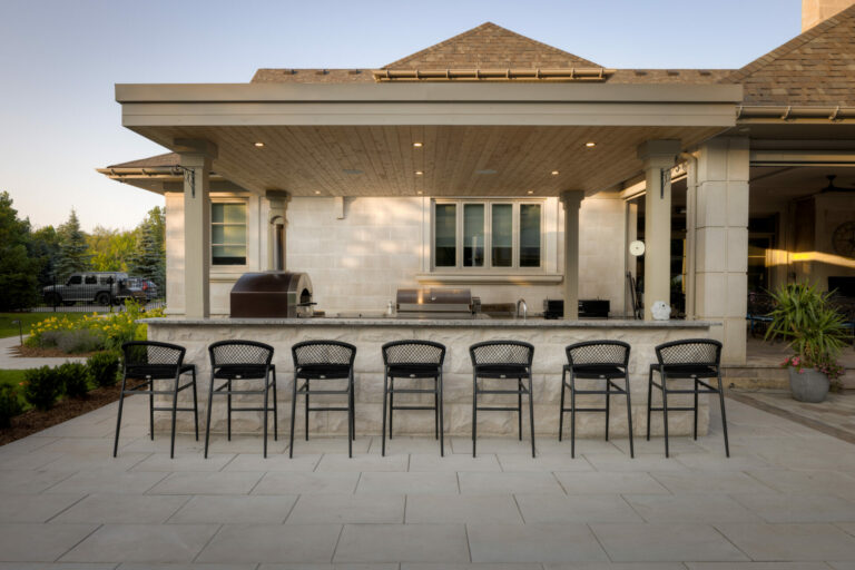 Outdoor patio kitchen with seven bar stools, brick oven, and covered pergola. Stylish design features ample seating and modern appliances, surrounded by greenery.