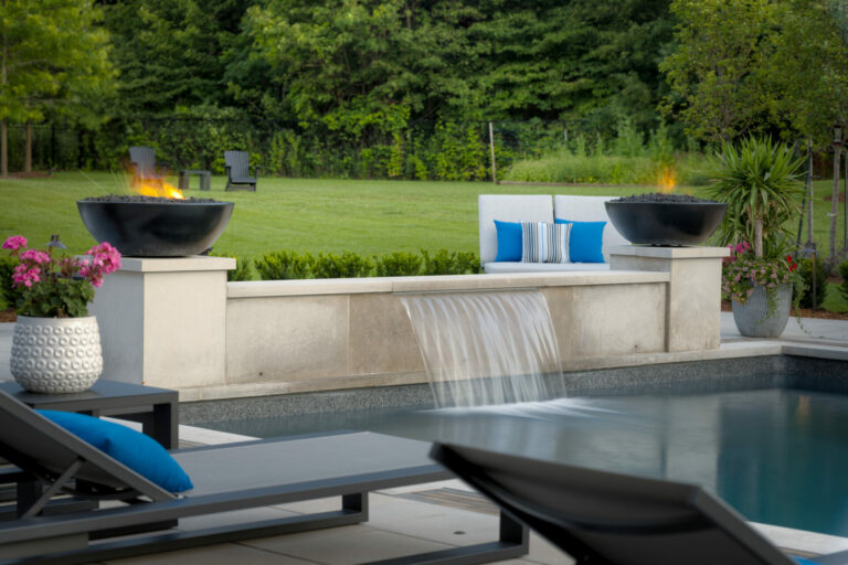 A modern backyard with a pool, waterfall, fire bowls, lounge chairs, and lush greenery, creating a serene and luxurious outdoor setting.