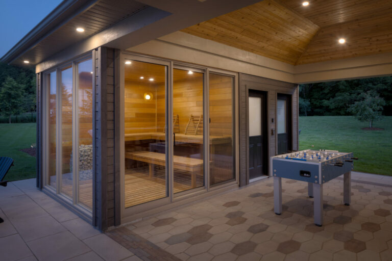 Modern sauna with glass walls, illuminated interior, and adjacent foosball table. Situated in a spacious backyard with a well-maintained lawn.