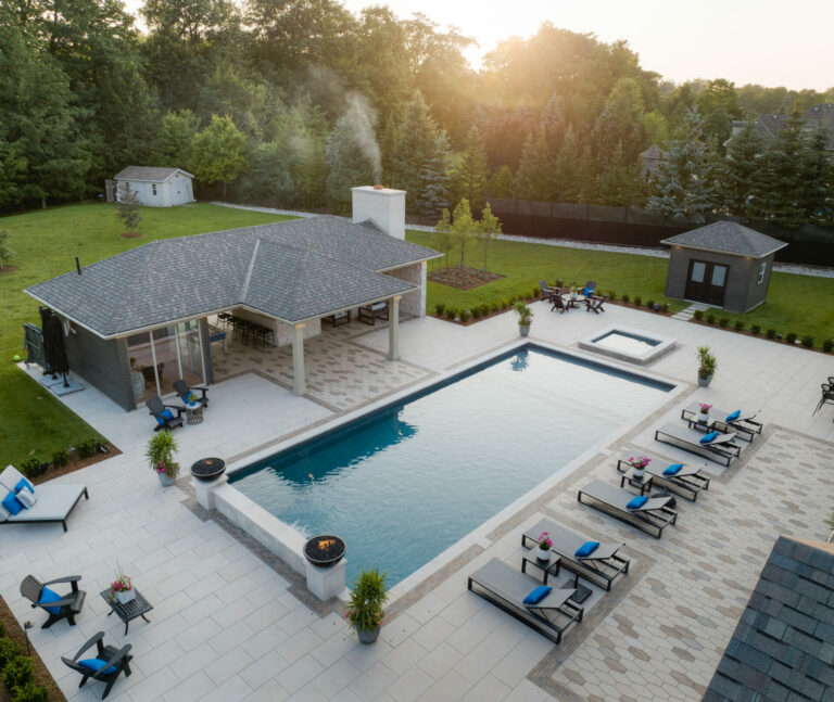 Spacious backyard with a large pool, lounge chairs, and a covered patio. Surrounded by lush greenery and two small buildings nearby.