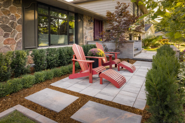 Two red Adirondack chairs on a stone patio, surrounded by greenery and a stone house with large windows. Peaceful outdoor scene.