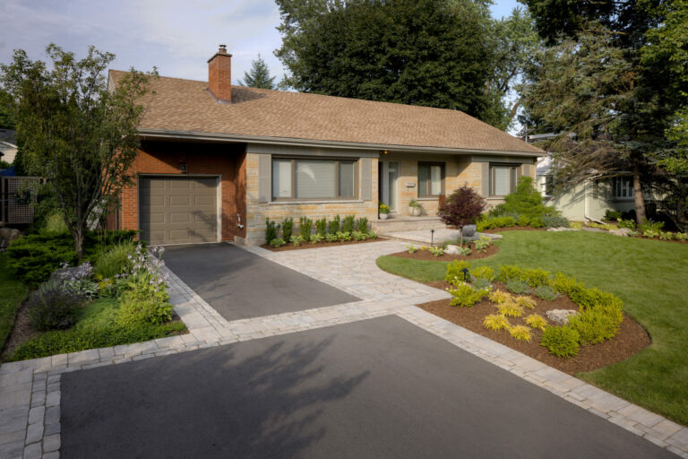 A well-maintained suburban home with a brick exterior, landscaped garden, and paved driveway, surrounded by trees and greenery on a sunny day.