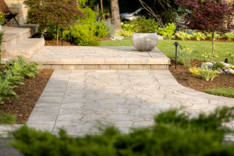 A well-manicured garden path with stone tiles, surrounded by greenery and small trees, leads to an elevated area with a large planter.