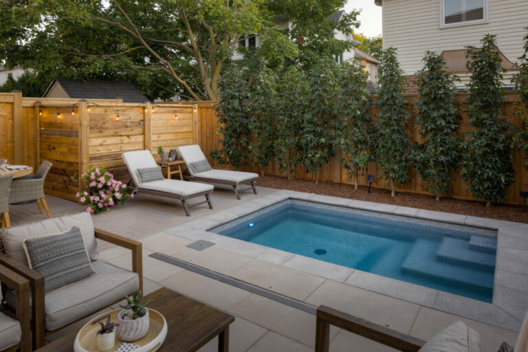 Cozy backyard scene with a small pool, lounge chairs, wooden fence, and greenery. Comfortable seating and lighting create an inviting outdoor atmosphere.